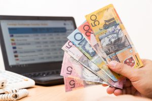 Hand holding Australian dollar in office with computer screen showing foreign exchange table in background