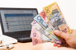 Hand holding Australian dollar in office with computer screen showing foreign exchange table in background